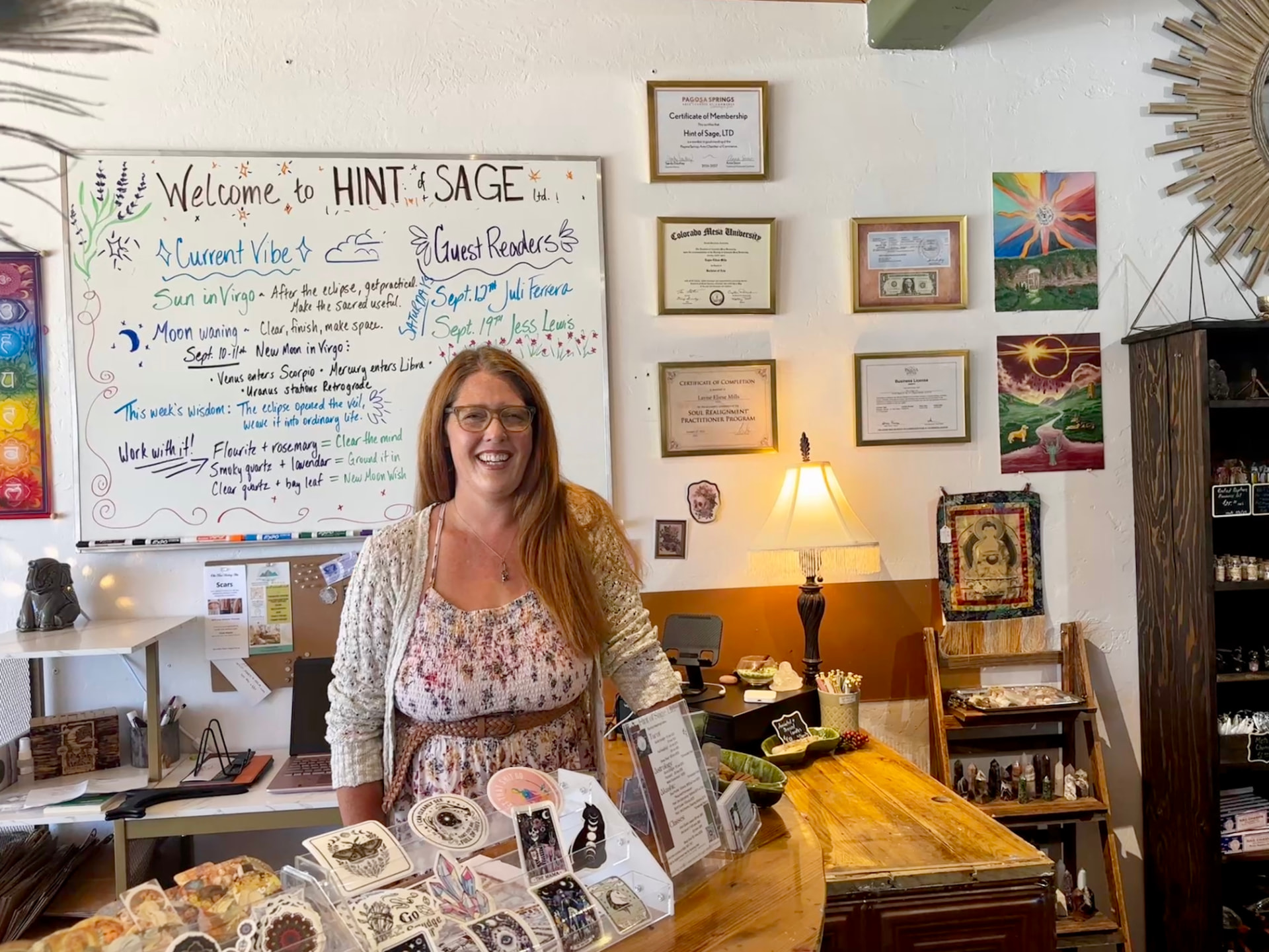 Layne smiling behind the counter at Hint of Sage, with the hand-lettered welcome board and framed certificates on the wall